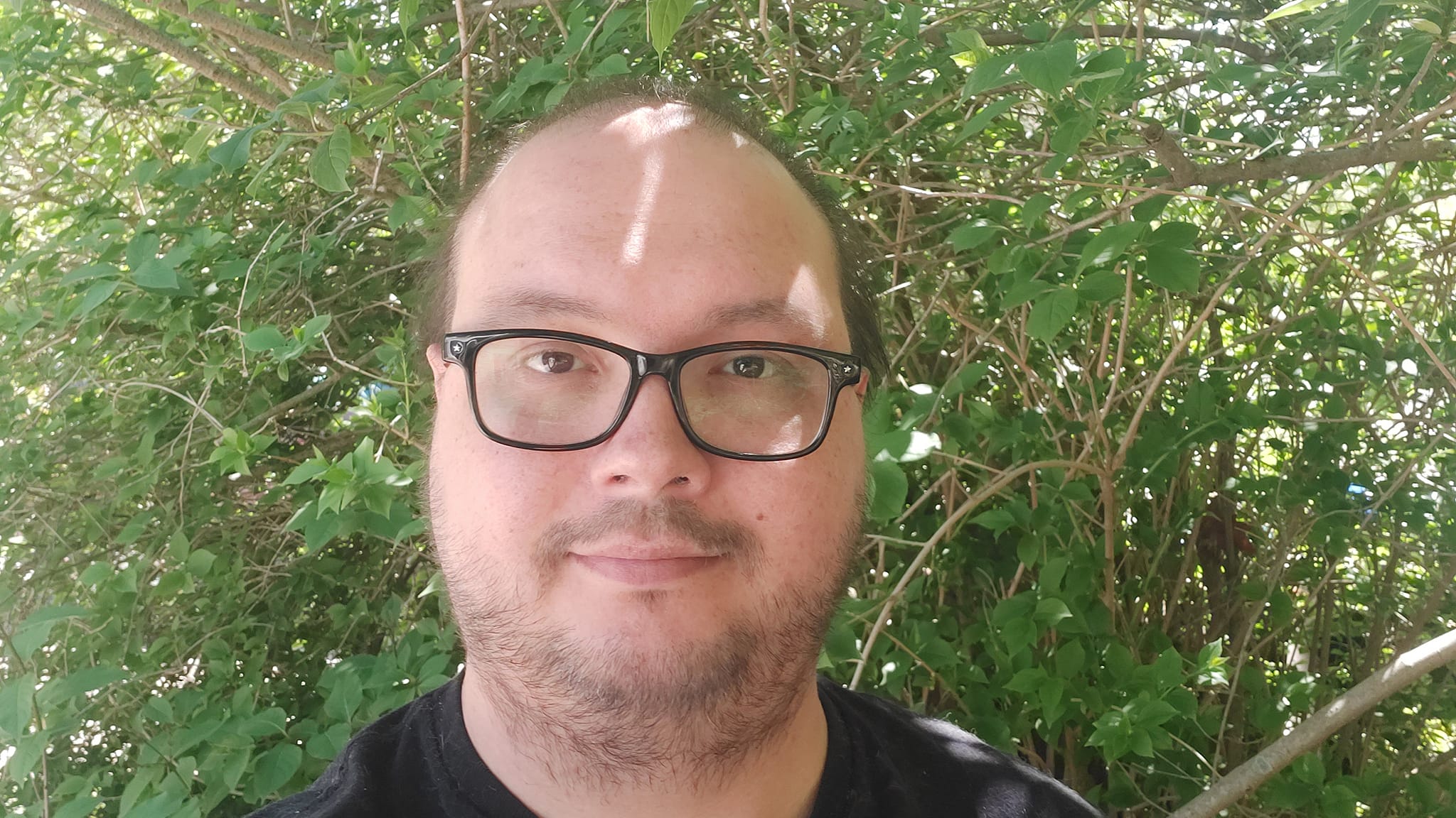 A man wearing black shirt stands in front of a bush full of green leaves, he is growing stubble